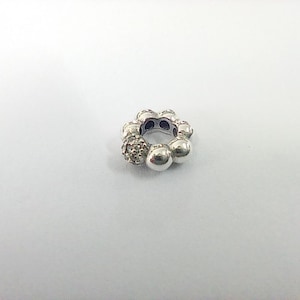 May include: A silver charm with a floral design. The charm features a ring of small, round, polished silver beads and a cluster of smaller, sparkling beads. The charm is designed to be worn on a bracelet or necklace.