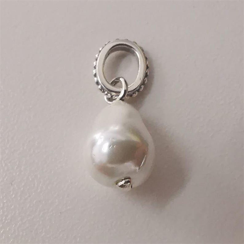 May include: A silver-toned charm with a large, pear-shaped white pearl pendant. The charm has a small, round, beaded top ring and a small loop connecting to the pearl. The pearl has a smooth, lustrous surface.