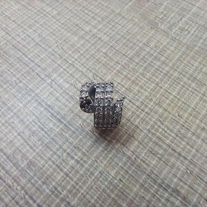 May include: A silver-toned, spiral-shaped earring with multiple rows of small, clear gemstones. The earring is set against a light brown, textured surface, showcasing its intricate design and sparkling details.