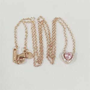 May include: A rose gold necklace with a heart-shaped pink gemstone pendant, surrounded by a halo of smaller clear stones. The chain is delicate and has a lobster clasp closure.