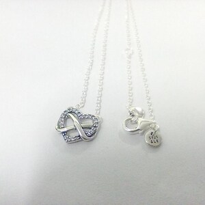 May include: A silver necklace with a heart-shaped pendant and an infinity symbol, set with small, clear gemstones. The necklace has a fine chain and a lobster clasp. The clasp has a small tag.