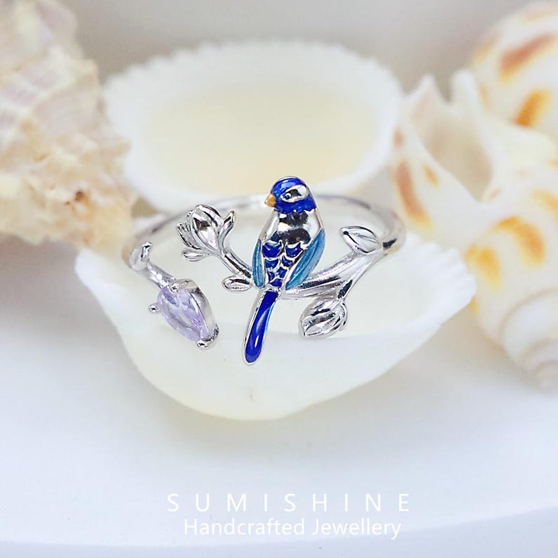 May include: A silver ring featuring a blue and silver bird perched on a branch with a purple gemstone. The bird has blue and silver enamel detailing. The ring is set against a white background with seashells.