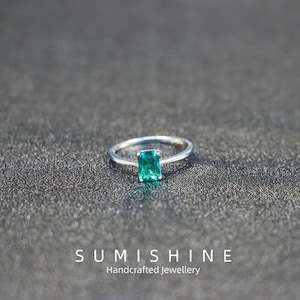Peut inclure: Bague en argent ornée d'une pierre verte rectangulaire taille émeraude. La bague est posée sur une surface texturée gris foncé. Le nom de la marque "SUMISHINE" et le texte "Handcrafted Jewellery" sont visibles.