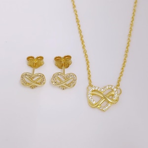 May include: A gold-colored jewelry set featuring a necklace and matching earrings. The necklace has a pendant in the shape of a heart intertwined with an infinity symbol, embellished with small, clear stones. The earrings mirror the pendant design.