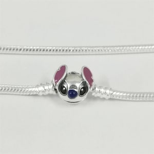 May include: A silver charm depicting a cartoon character's face with pink ears, black eyes, and a blue nose. The charm is on a silver chain, suitable for a bracelet or necklace. The background is plain white.