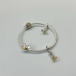 May include: Silver bracelet with several charms. The bracelet includes a Mickey Mouse charm, a carriage charm, and a character charm with yellow detailing. The bracelet is set against a white backdrop.