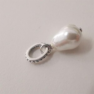 May include: A pear-shaped, white pearl charm suspended from a silver oval link. The link is marked with "925 ALE" and has a beaded edge. The pearl has a smooth, lustrous surface.