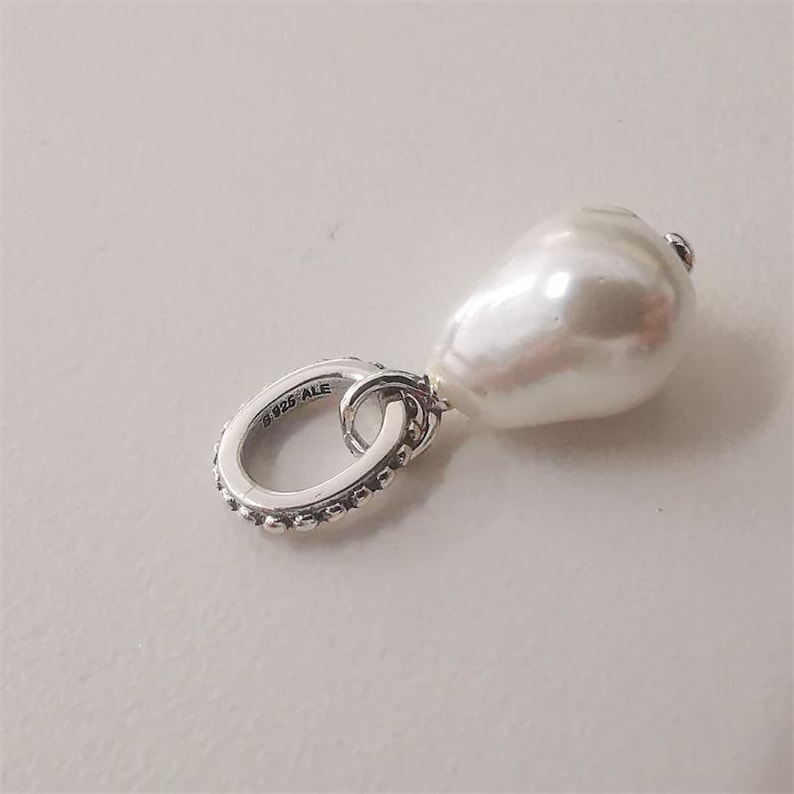May include: A pear-shaped, white pearl charm suspended from a silver oval link. The link is marked with "925 ALE" and has a beaded edge. The pearl has a smooth, lustrous surface.