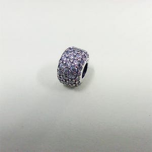 May include: A silver bead charm, covered in small, clear crystals. The charm is cylindrical with a rounded shape and a dark hole on one side. The crystals give the charm a sparkling appearance.