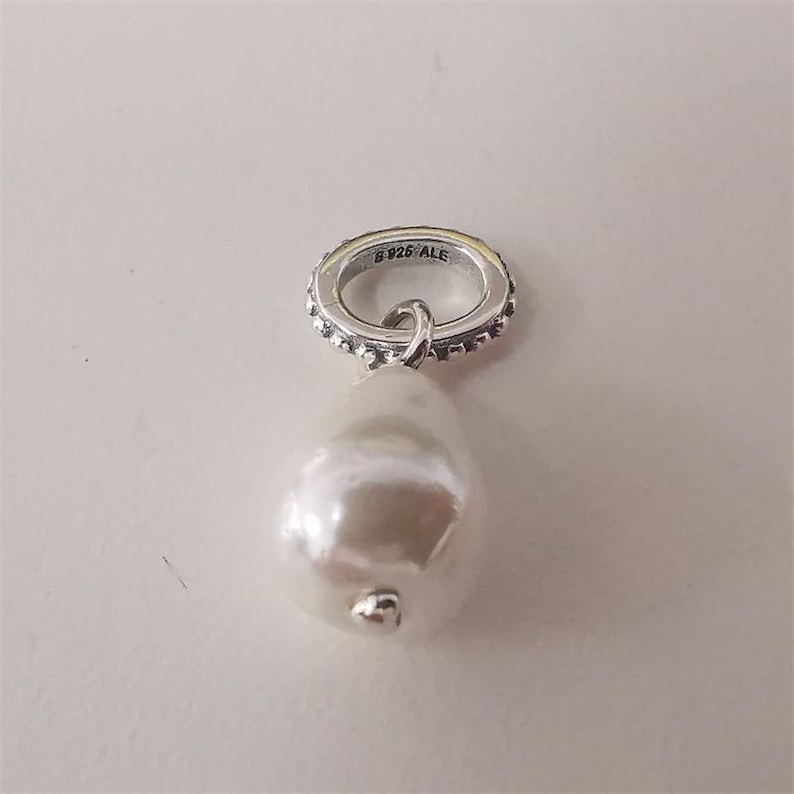 May include: A silver charm with a large, pear-shaped white pearl pendant. The charm's ring is adorned with small, sparkling accents and the text "S 925 ALE". The pearl has a smooth, lustrous surface and a small silver accent.