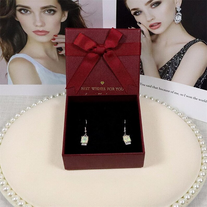 May include: A burgundy gift box with a matching bow, open to reveal a pair of square, light-colored gemstone earrings. The box has the text "BEST WISHES FOR YOU" printed on it. The earrings are displayed on a white surface.