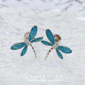 May include: A pair of dragonfly stud earrings. Each earring features a silver-toned body and wings in a teal enamel finish. A small, clear gemstone is set at the center of each dragonfly. The earrings are displayed on a textured white surface.