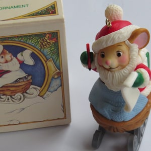 May include: A white mouse wearing a red Santa hat and a green and red Santa suit is riding in a brown sleigh. The mouse is holding a red pencil and a white piece of paper. The sleigh has two wheels and is on a white background.