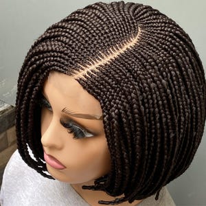 May include: A dark brown, braided bob wig. The wig features a lace front and a center part. The braids are tightly woven and create a textured appearance. The wig is styled on a mannequin head, showcasing the shape and style of the wig.