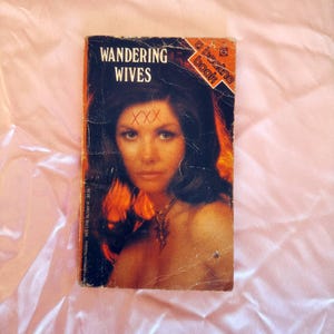 May include: Vintage paperback book titled "Wandering Wives" with a woman's portrait on the cover. The cover is predominantly black with orange and red accents. The woman has dark hair and a necklace. The book is a "a bee-line book".