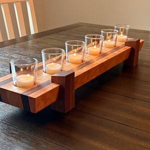 Może przedstawiać: Drewniany świecznik z sześcioma szklanymi świecznikami wotywnymi. Uchwyt wykonany jest z naprzemiennych pasków jasnego i ciemnego drewna. Każdy wotyw zawiera zapaloną świecę, rzucającą ciepłą poświatę. Świecznik stoi na ciemnym drewnianym stole.