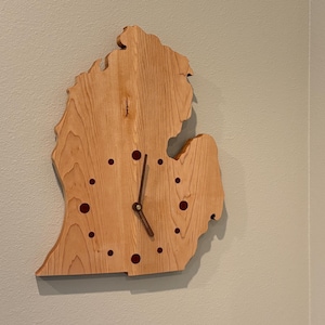May include: A wooden clock in the shape of the state of Michigan. The clock has a light brown finish with dark red dots marking the hours. The clock hands are dark brown.