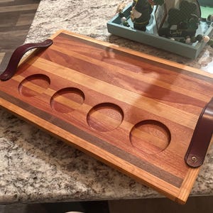 May include: A rectangular wooden serving tray with four circular cutouts for glasses. The tray features alternating stripes of light and dark wood, with dark brown leather handles on each end. The tray is designed for serving drinks or snacks.