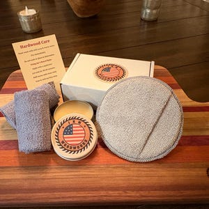 May include: A hardwood care kit on a wooden cutting board. The kit includes a box, a tin with an American flag design, a round gray pad, two gray cloths, and a small jar of wax. A card with care instructions is also included.
