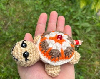May include: A crocheted turtle keychain with a beige head and limbs, and a shell featuring orange, brown, and white patterns. The turtle has black bead eyes and a red bead on its back. A small red and orange bead is attached to the keychain.
