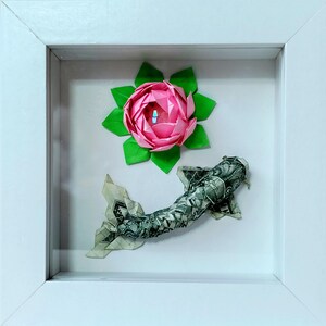 Pode incluir: Obra de arte de origami emoldurada. Apresenta uma flor de lótus rosa com folhas verdes e um peixe feito com uma nota de dólar. A obra está sobre um fundo branco dentro de uma moldura branca.
