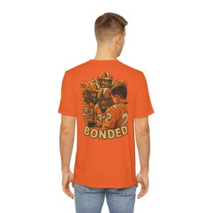 May include: Orange t-shirt featuring a graphic of a football team huddle with the word "BONDED" in a vintage style. The players wear orange and white uniforms and helmets. The shirt is a solid orange color.