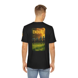 May include: Black t-shirt featuring a rectangular graphic on the back with the word "ENDURE" in gold letters. The graphic depicts a football team running on a green field with a sunset background. The shirt is made of a soft, comfortable material.