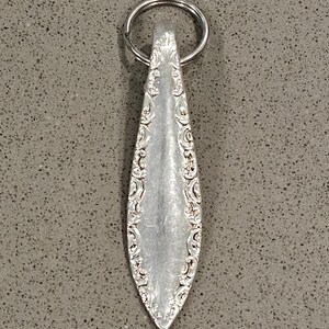 May include: Silver spoon keychain with ornate floral detailing along the edges. The spoon has a teardrop shape and is attached to a silver key ring. The background is a speckled gray surface.