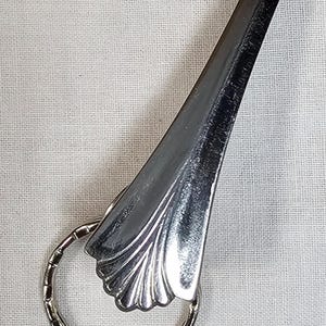May include: A silver-colored key ring with a decorative, fan-shaped top. The key ring is attached to a long, tapered piece of metal. The key ring is round with a textured surface. The background is a light-colored fabric.