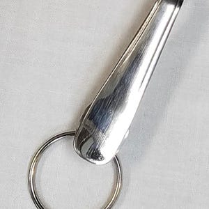 May include: A shiny, silver-colored key holder with a ring and a streamlined, elongated body. The key holder is made of metal and has a polished finish. The ring is circular and attached to the body. The background is a white, textured fabric.
