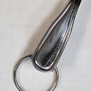 May include: A silver-colored keychain with a spoon-shaped top and a round key ring. The spoon-shaped top has a smooth, reflective surface. The key ring is made of a similar silver-colored metal.