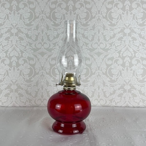 May include: A vintage oil lamp with a ruby red glass base and a clear glass chimney. The lamp has a brass burner and a round, bulbous shape. The lamp is designed to sit on a table or other flat surface.