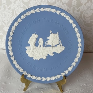 May include: A blue decorative plate with a white relief design commemorating the Apollo 11 moon landing. The plate features the text "MAN ON THE MOON" and "APOLLO ELEVEN JULY 20 1969". A white laurel wreath design borders the plate.