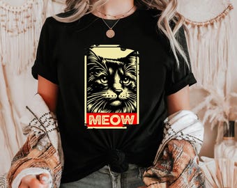 Puede incluir: Camiseta negra con un diseño rectangular que presenta la cara de un gato y la palabra "MEOW" en rojo. El gato es negro con detalles blancos, sobre un fondo amarillo. Ilustración gráfica.