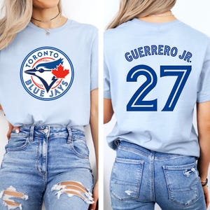Puede incluir: Camiseta azul claro con el logotipo de los Blue Jays de Toronto en la parte delantera y "GUERRERO JR. 27" en la parte trasera. El logotipo incluye un arrendajo azul, una hoja de arce y las palabras "TORONTO BLUE JAYS". El número 27 es azul marino.