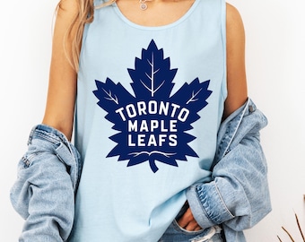May include: Light blue tank top featuring a large navy blue maple leaf graphic with the text "TORONTO MAPLE LEAFS" in white. The tank top is sleeveless and made of a soft material.