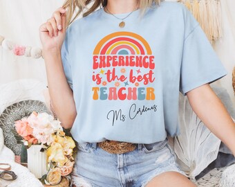 May include: Light blue t-shirt with a rainbow graphic and the text "EXPERIENCE is the best TEACHER Ms. Cardenas" in red, orange, and blue. The shirt is made of a soft material and has short sleeves.