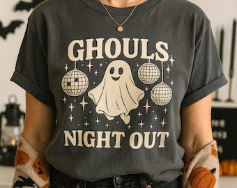 Puede incluir: Camiseta gris oscuro con el texto "GHOULS NIGHT OUT" y un gráfico de fantasma. La camiseta presenta un fantasma, bolas de discoteca y estrellas. La persona lleva un suéter beige con calabazas naranjas y murciélagos negros.
