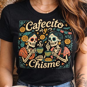 A woman wearing a black Latina graphic tee featuring the vibrant, vintage style illustration for 'Cafecito y Chisme' (Coffee and Gossip). The design shows two skeletons (calaveras) sharing a cup of coffee and eating pan dulce in a detailed Mexican folk art style with Dia de los Muertos (Day of the Dead) floral accents. The lifestyle setting includes a real concha and latte on a rustic table, highlighting a cozy Spanglish apparel aesthetic.