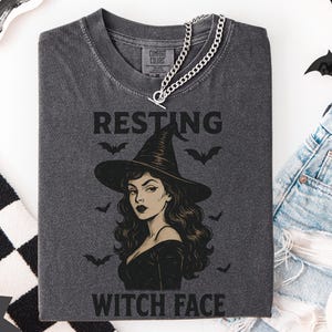 May include: A graphite-coloured t-shirt featuring a witch graphic and the words "RESTING WITCH FACE." The image includes a silver chain necklace, bat decorations, and denim shorts, creating a Halloween theme.
