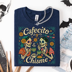 Cafecito y Chisme Shirt, Womens Funny Mexican Shirt for Latina, Halloween Skeleton Dia De Los Muertos Tshirt, Gift for Mom Abuelita — black Comfort Colors–style tee with two calavera amigas sipping cafecito, flowers, papel picado and concha graphic; folded flat lay with ripped black jeans, fall leaves, bat cutout and skull prop; cozy Latina coffee graphic tee for spooky season.