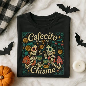 Cafecito y Chisme Shirt, Womens Funny Mexican Shirt for Latina, Halloween Skeleton Dia De Los Muertos Tshirt, Gift for Mom Abuelita — black Comfort Colors–style tee with two calavera amigas sipping cafecito, flowers, papel picado and concha graphic; folded flat lay with ripped black jeans, fall leaves, bat cutout and skull prop; cozy Latina coffee graphic tee for spooky season.