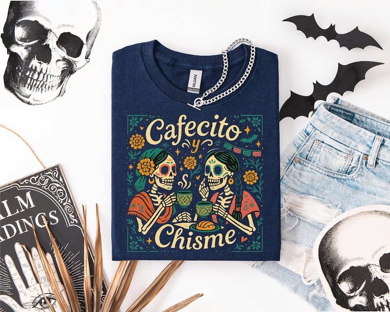 Cafecito y Chisme Shirt, Womens Funny Mexican Shirt for Latina, Halloween Skeleton Dia De Los Muertos Tshirt, Gift for Mom Abuelita — black Comfort Colors–style tee with two calavera amigas sipping cafecito, flowers, papel picado and concha graphic; folded flat lay with ripped black jeans, fall leaves, bat cutout and skull prop; cozy Latina coffee graphic tee for spooky season.