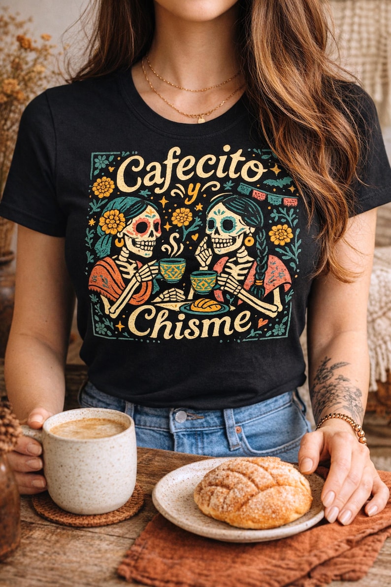 A woman wearing a black Latina graphic tee featuring the vibrant, vintage style illustration for 'Cafecito y Chisme' (Coffee and Gossip). The design shows two skeletons (calaveras) sharing a cup of coffee and eating pan dulce in a detailed Mexican folk art style with Dia de los Muertos (Day of the Dead) floral accents. The lifestyle setting includes a real concha and latte on a rustic table, highlighting a cozy Spanglish apparel aesthetic.