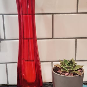 May include: A tall, vibrant red glass vase with a flared top. The vase is next to a small succulent in a gray and copper-toned pot, resting on a book titled "The Secret Panel". The background features white tiles.