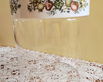 Pode incluir: Um frasco de vidro transparente com uma borda branca com um design de frutas colorido. O frasco é cilíndrico e assenta sobre uma toalha de renda branca. As ilustrações de frutas incluem limões, tomates e outros produtos.