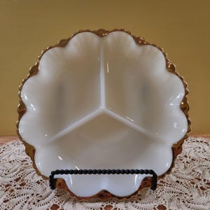 May include: A white, divided serving dish with a scalloped edge and gold trim. The dish is divided into three sections and is displayed on a black metal stand. The dish is likely vintage and suitable for serving appetizers or snacks.