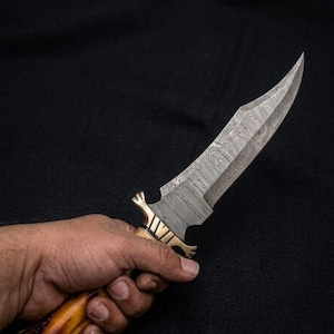 May include: A handcrafted knife with a patterned steel blade and a bone handle. The knife features a brass bolster and a curved blade. The handle is a light brown color. The knife is held against a black background.