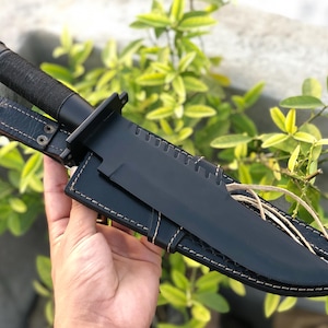 May include: A black survival knife with a textured handle and a leather sheath. The knife blade is black and has a straight edge. The sheath is black leather with white stitching.
