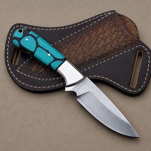May include: A fixed-blade knife with a silver steel blade and a handle featuring a bright turquoise and black geometric pattern. The knife is positioned on top of a dark brown leather sheath with a woven texture and white stitching along the edges.